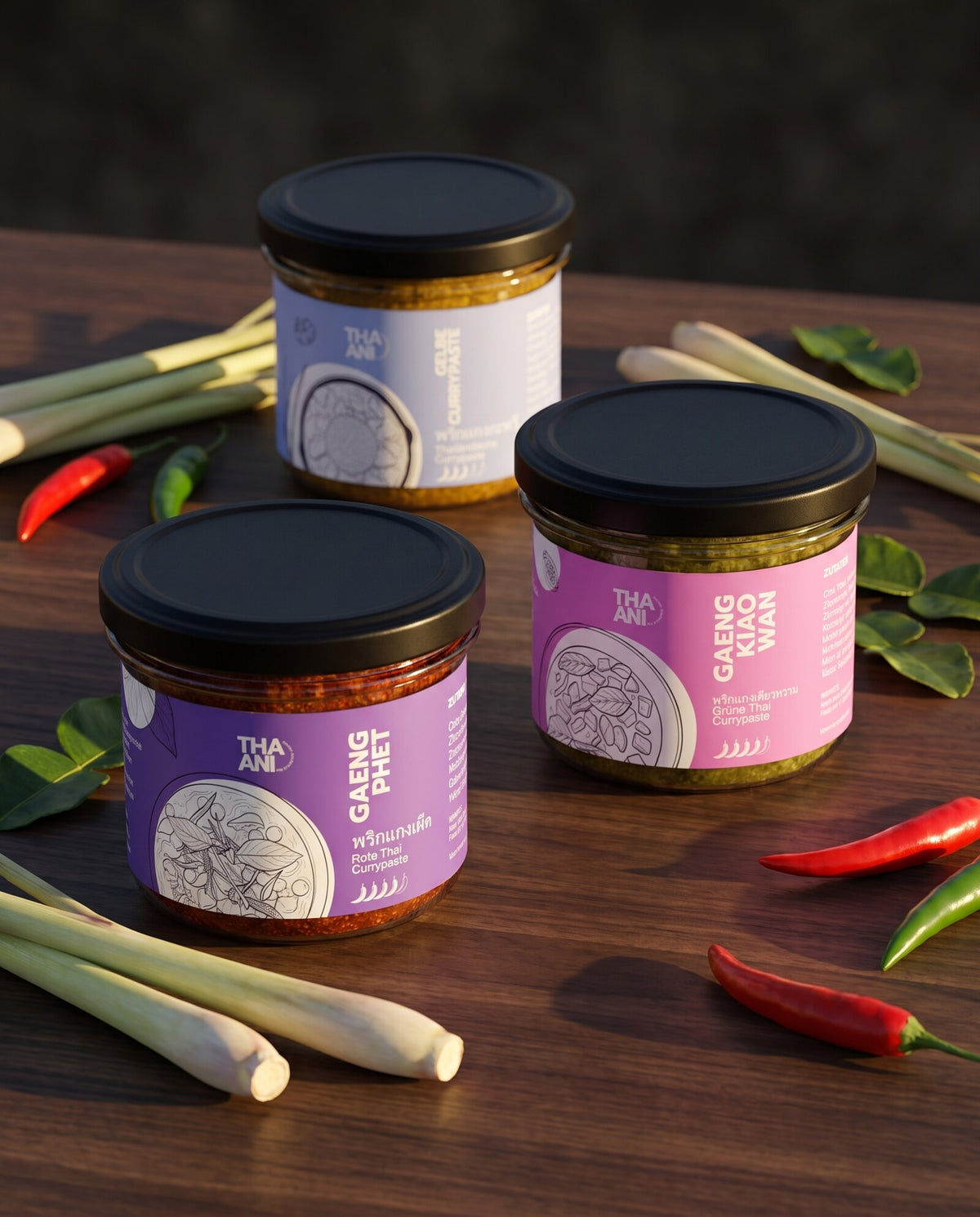 Drei THAANI-Gläser mit Currypaste auf einem Holztisch, daneben Zitronengras, Chilischoten und Kaffirlimettenblätter
